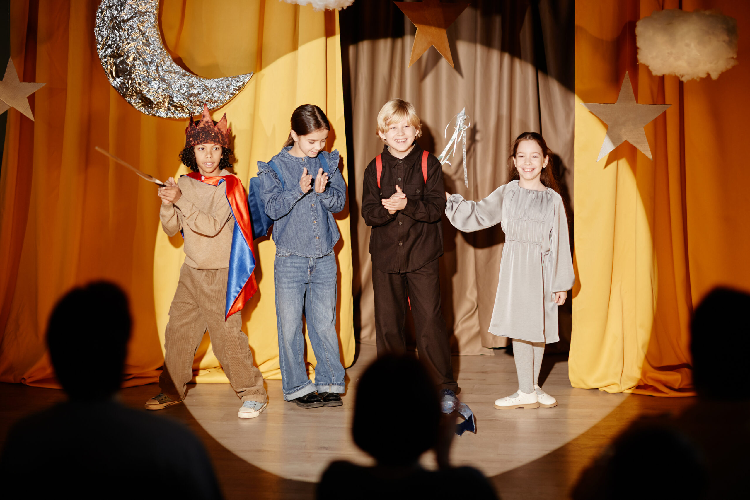Group of children dressed in costumes performing a play on stage with bright yellow curtains in background creating a lively atmosphere with stars, moon props surrounding them